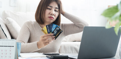 Concerned woman holding several credit cards while looking at a laptop, with a calculator, notebook, and calendar on the table.