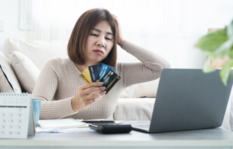Concerned woman holding several credit cards while looking at a laptop, with a calculator, notebook, and calendar on the table.