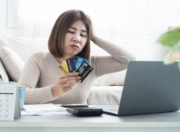 Concerned woman holding several credit cards while looking at a laptop, with a calculator, notebook, and calendar on the table.