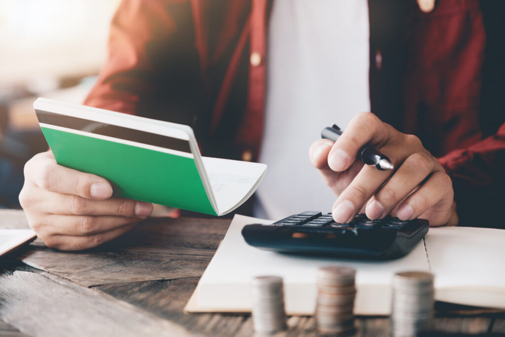 Person using a calculator while holding a passbook, with stacked coins on a table, representing budgeting or personal finance.