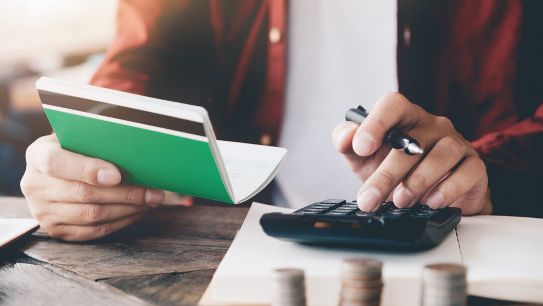 Person using a calculator while holding a passbook, with stacked coins on a table, representing budgeting or personal finance.