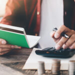Person using a calculator while holding a passbook, with stacked coins on a table, representing budgeting or personal finance.