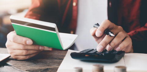 Person using a calculator while holding a passbook, with stacked coins on a table, representing budgeting or personal finance.