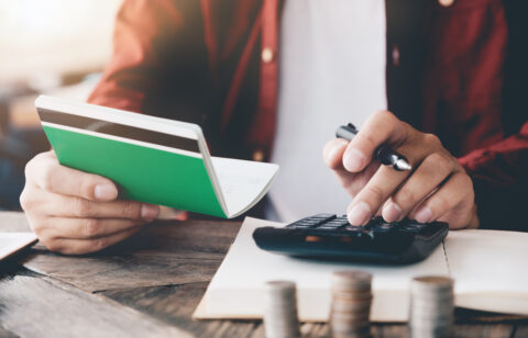 Person using a calculator while holding a passbook, with stacked coins on a table, representing budgeting or personal finance.
