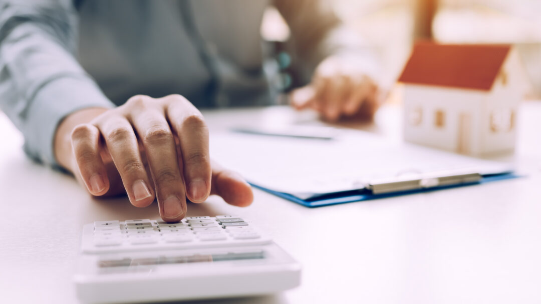 Person using a calculator beside a miniature house model and mortgage paperwork.