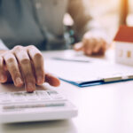 Person using a calculator beside a miniature house model and mortgage paperwork.