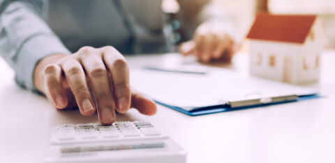 Person using a calculator beside a miniature house model and mortgage paperwork.