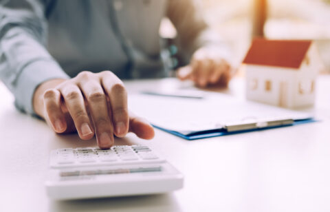Person using a calculator beside a miniature house model and mortgage paperwork.