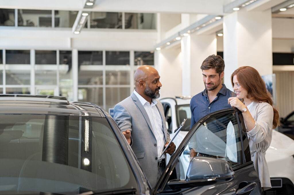 Car salesperson speaking with a couple beside a vehicle in a dealership showroom, discussing features before a purchase.