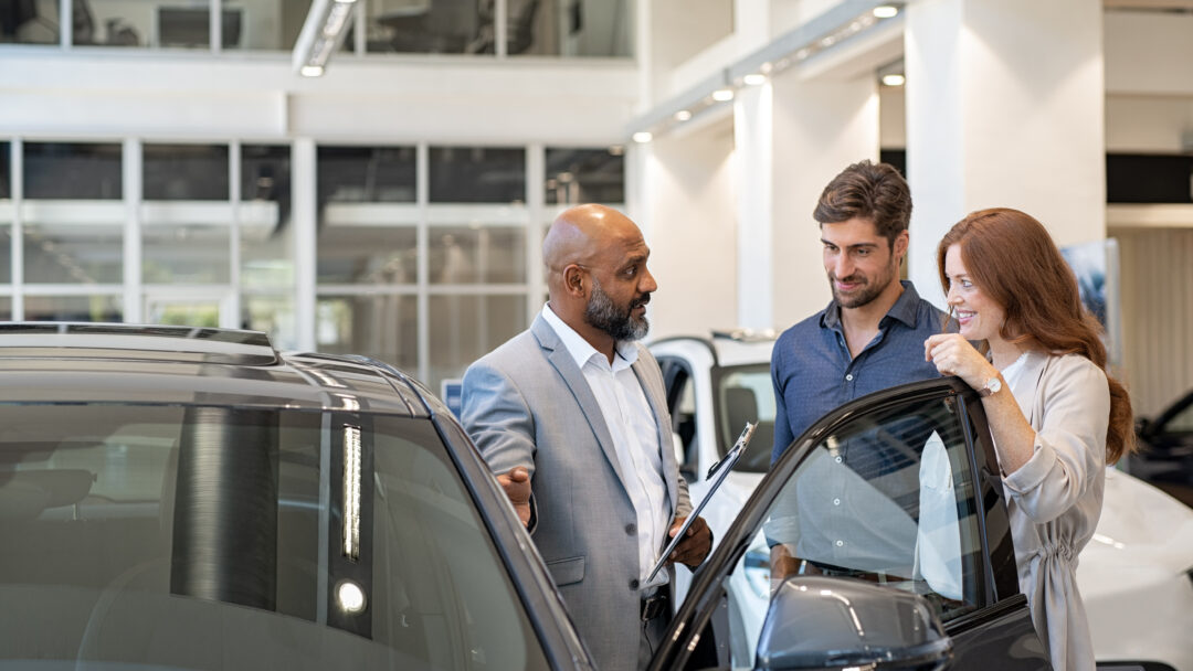 Car salesperson speaking with a couple beside a vehicle in a dealership showroom, discussing features before a purchase.