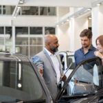 Car salesperson speaking with a couple beside a vehicle in a dealership showroom, discussing features before a purchase.