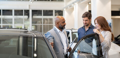 Car salesperson speaking with a couple beside a vehicle in a dealership showroom, discussing features before a purchase.