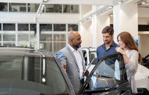 Car salesperson speaking with a couple beside a vehicle in a dealership showroom, discussing features before a purchase.