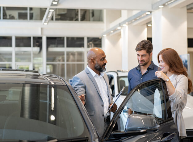 Car salesperson speaking with a couple beside a vehicle in a dealership showroom, discussing features before a purchase.