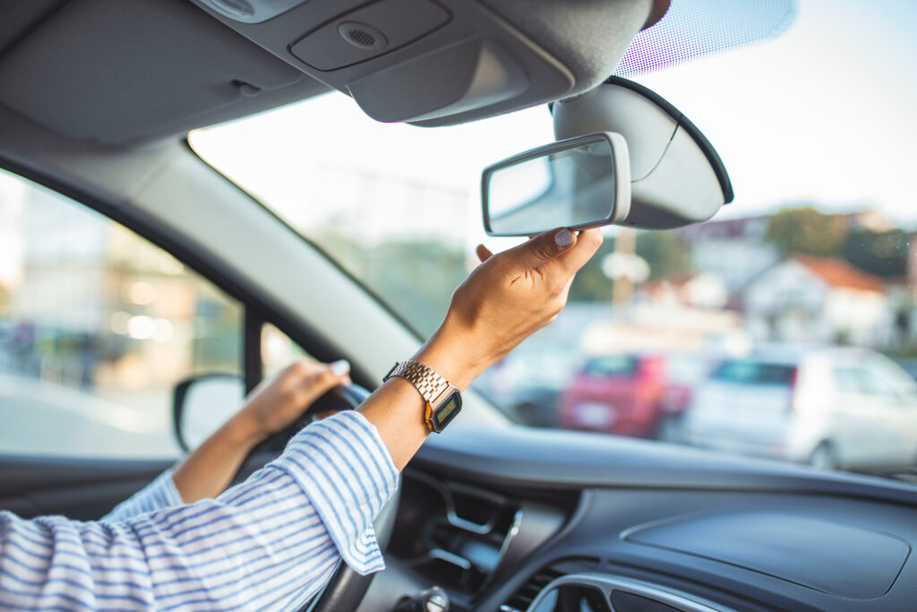 Driver adjusting the rearview mirror inside a car before driving, with one hand on the steering wheel.