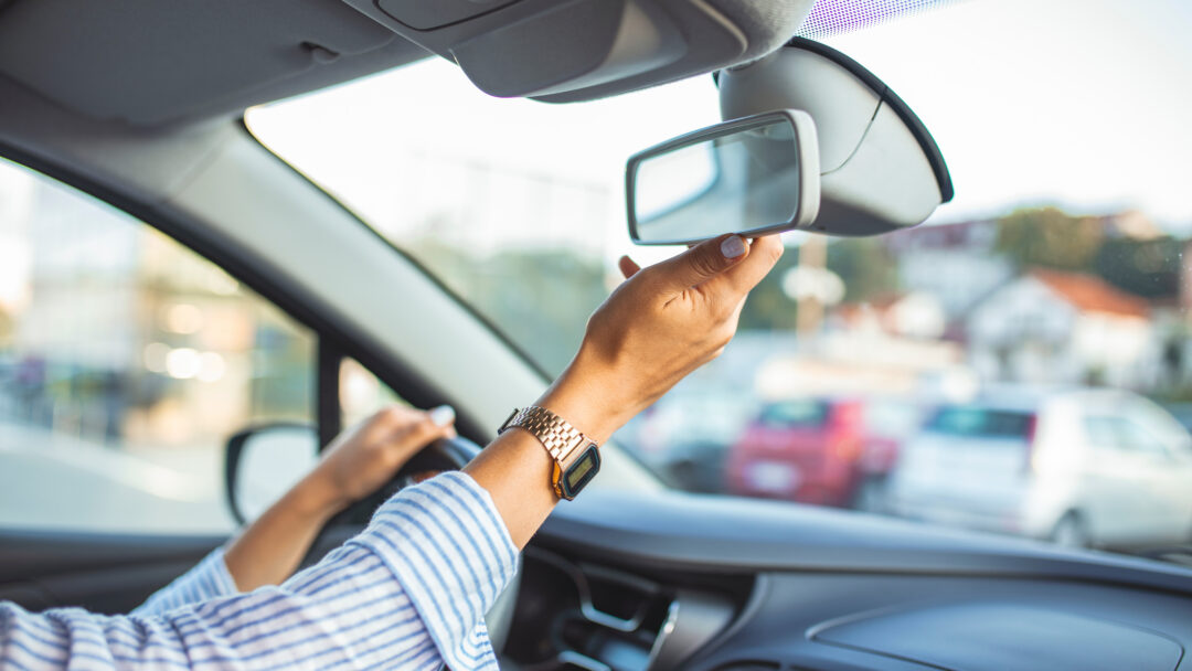 Driver adjusting the rearview mirror inside a car before driving, with one hand on the steering wheel.