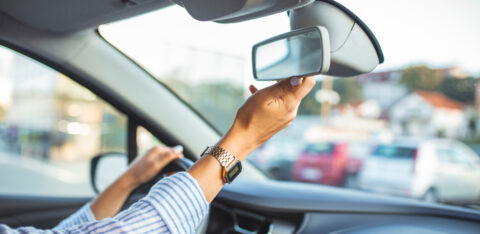 Driver adjusting the rearview mirror inside a car before driving, with one hand on the steering wheel.