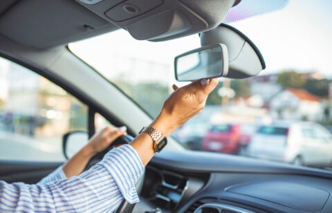 Driver adjusting the rearview mirror inside a car before driving, with one hand on the steering wheel.