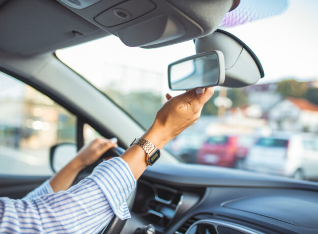 Driver adjusting the rearview mirror inside a car before driving, with one hand on the steering wheel.