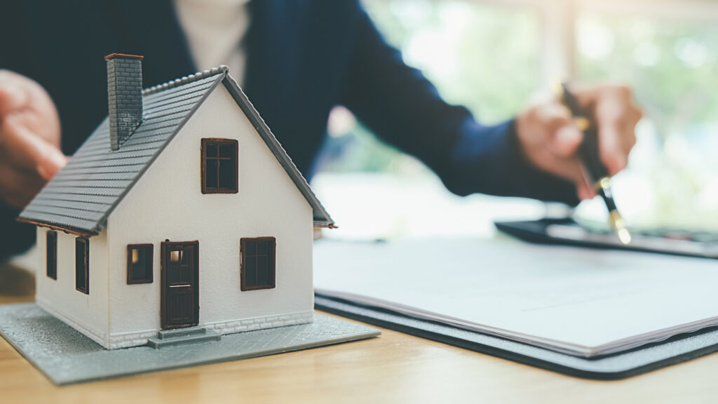 Model house on a desk beside paperwork, with a person signing documents in the background.