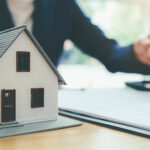 Model house on a desk beside paperwork, with a person signing documents in the background.