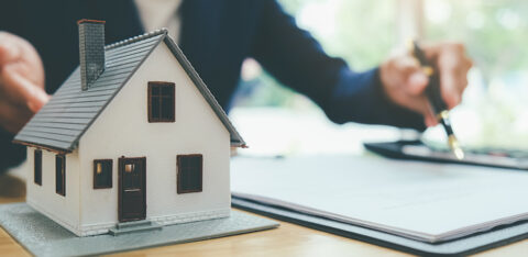 Model house on a desk beside paperwork, with a person signing documents in the background.