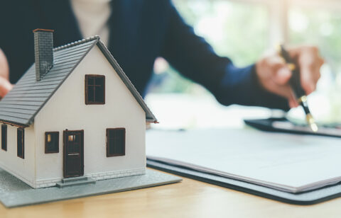 Model house on a desk beside paperwork, with a person signing documents in the background.