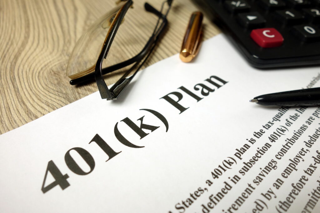 Close-up of a 401(k) retirement plan document with a calculator, eyeglasses, and pens on a desk.