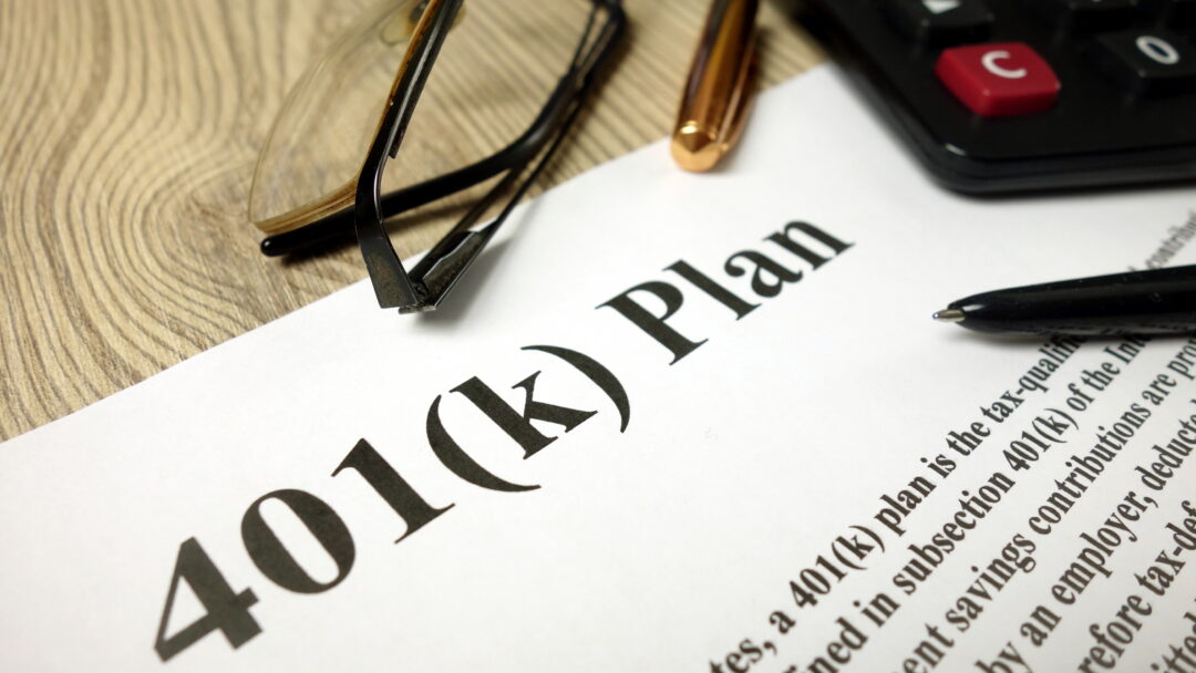 Close-up of a 401(k) retirement plan document with a calculator, eyeglasses, and pens on a desk.