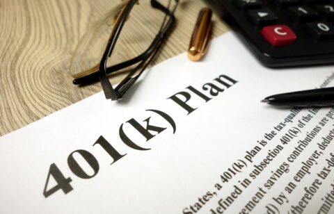 Close-up of a 401(k) retirement plan document with a calculator, eyeglasses, and pens on a desk.