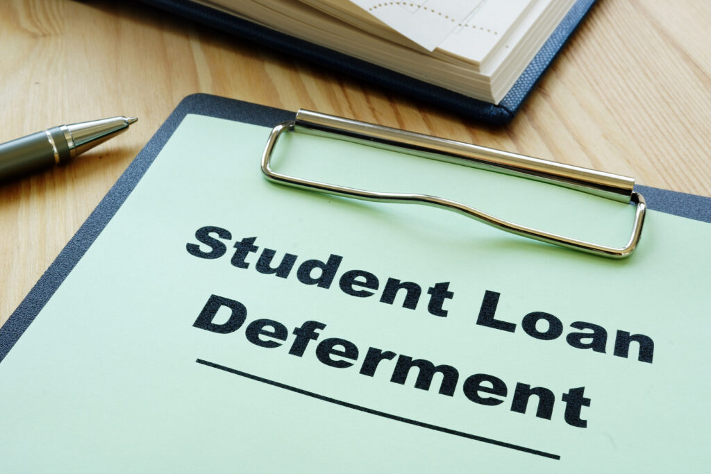 Clipboard labeled "Student Loan Deferment" on a desk with a pen and notebook.