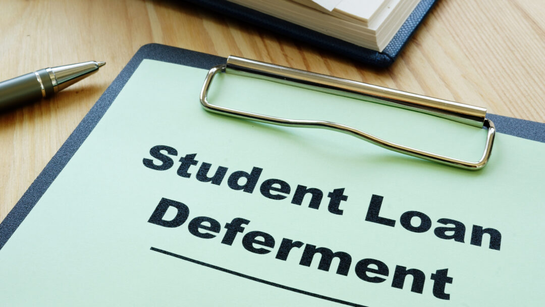 Clipboard labeled "Student Loan Deferment" on a desk with a pen and notebook.