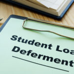 Clipboard labeled "Student Loan Deferment" on a desk with a pen and notebook.