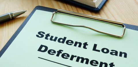 Clipboard labeled "Student Loan Deferment" on a desk with a pen and notebook.
