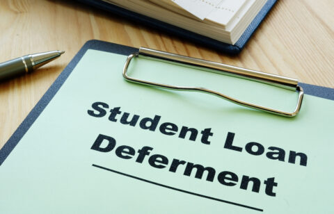 Clipboard labeled "Student Loan Deferment" on a desk with a pen and notebook.