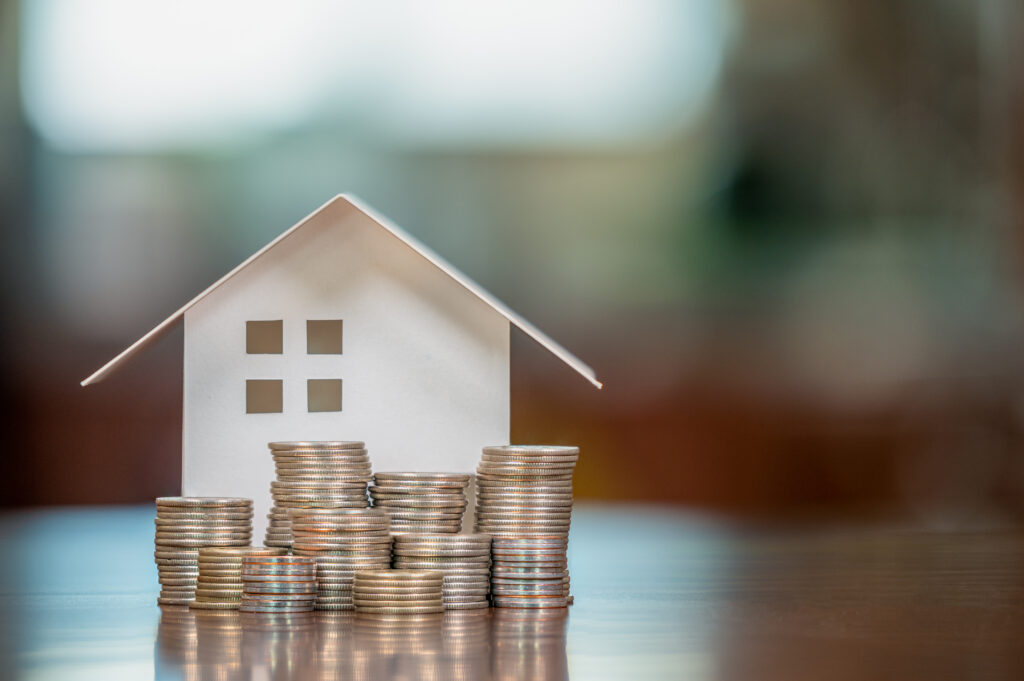 Paper house model surrounded by stacks of coins on a table, representing homeownership, savings, or mortgage planning.