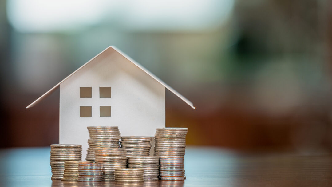 Paper house model surrounded by stacks of coins on a table, representing homeownership, savings, or mortgage planning.
