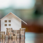 Paper house model surrounded by stacks of coins on a table, representing homeownership, savings, or mortgage planning.