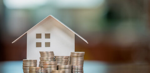 Paper house model surrounded by stacks of coins on a table, representing homeownership, savings, or mortgage planning.