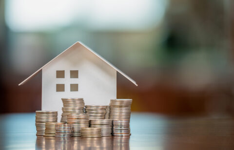 Paper house model surrounded by stacks of coins on a table, representing homeownership, savings, or mortgage planning.