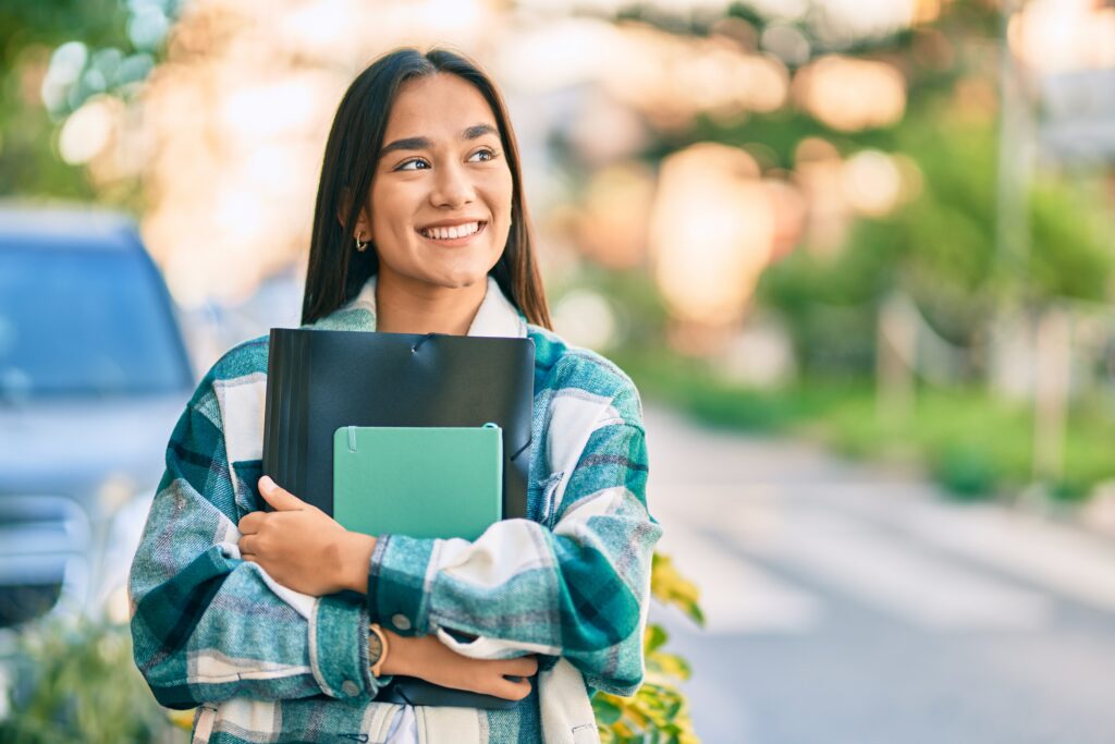 Smiling female college student holding notebooks and a folder while walking outdoors on campus.