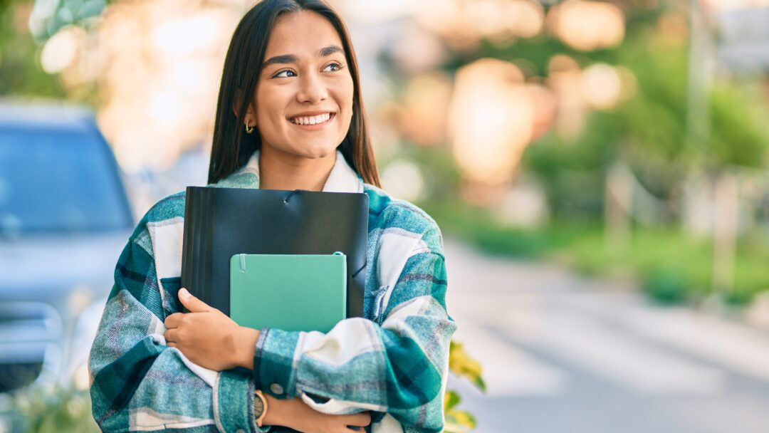 Smiling female college student holding notebooks and a folder while walking outdoors on campus.