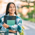Smiling female college student holding notebooks and a folder while walking outdoors on campus.