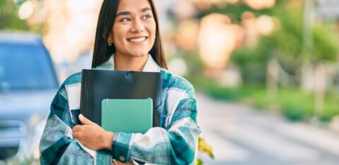 Smiling female college student holding notebooks and a folder while walking outdoors on campus.