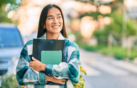 Smiling female college student holding notebooks and a folder while walking outdoors on campus.