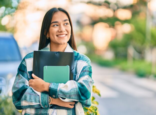 Smiling female college student holding notebooks and a folder while walking outdoors on campus.