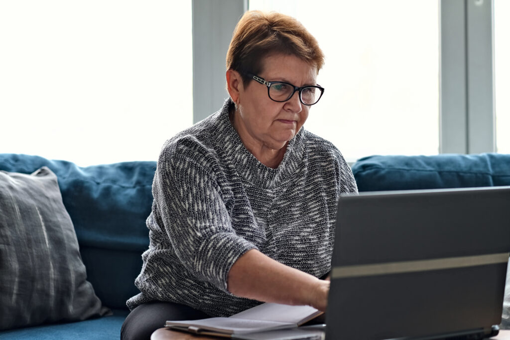 Older woman using a laptop at home while reviewing paperwork and taking notes, representing budgeting or personal finance management.
