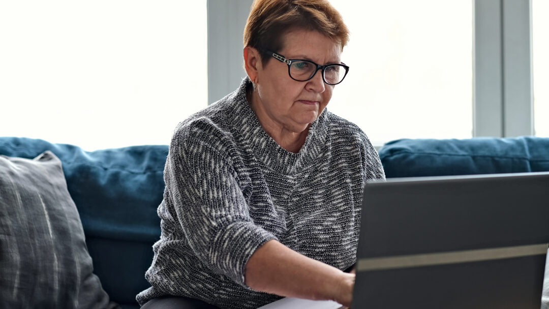Older woman using a laptop at home while reviewing paperwork and taking notes, representing budgeting or personal finance management.