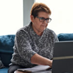 Older woman using a laptop at home while reviewing paperwork and taking notes, representing budgeting or personal finance management.