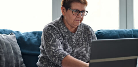 Older woman using a laptop at home while reviewing paperwork and taking notes, representing budgeting or personal finance management.